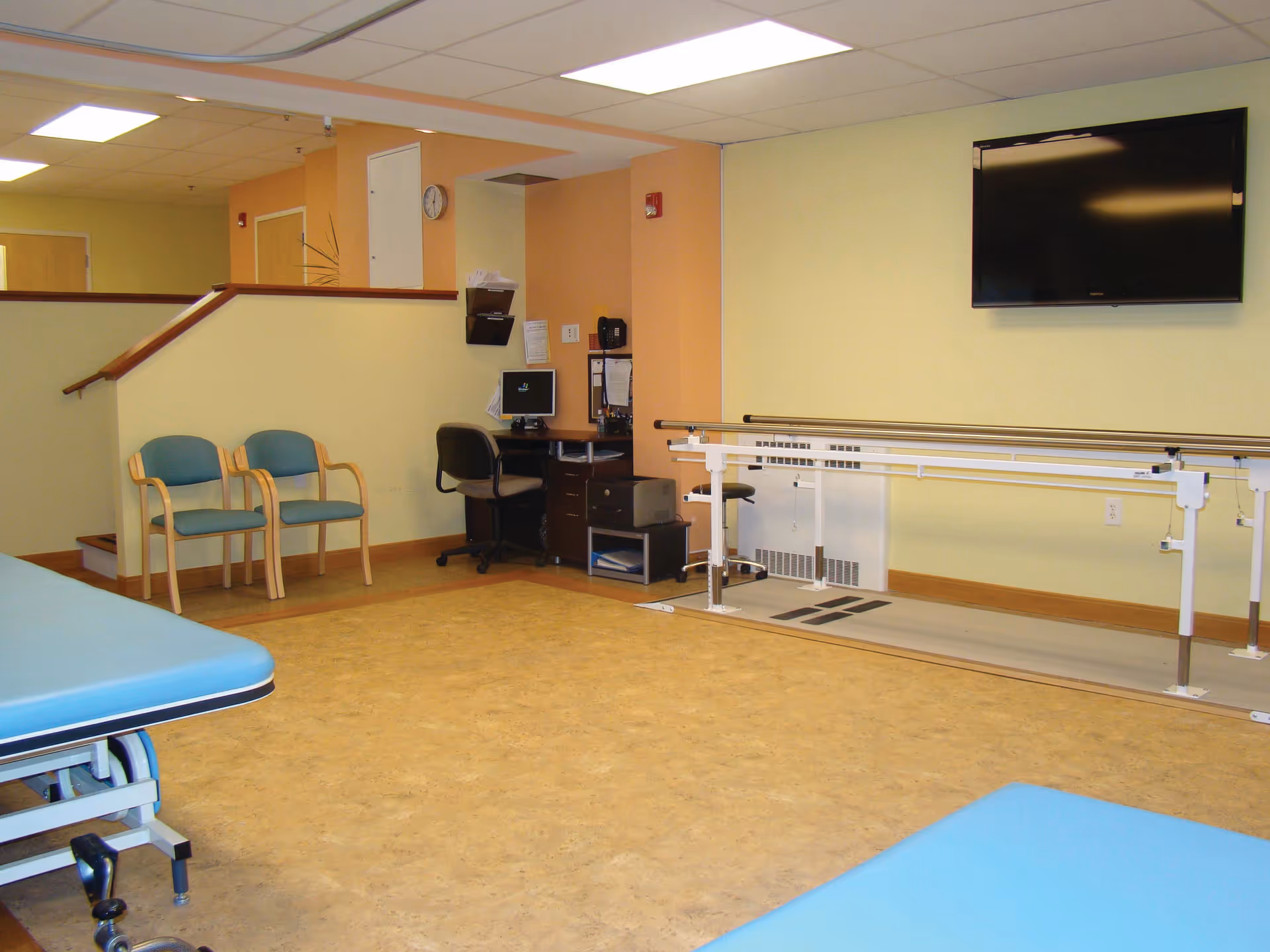 Interior of a rehabilitation room with parallel bars for walking exercises, two blue padded treatment tables, two blue chairs, a desk with a computer and office chair, and a wall-mounted flat screen TV.