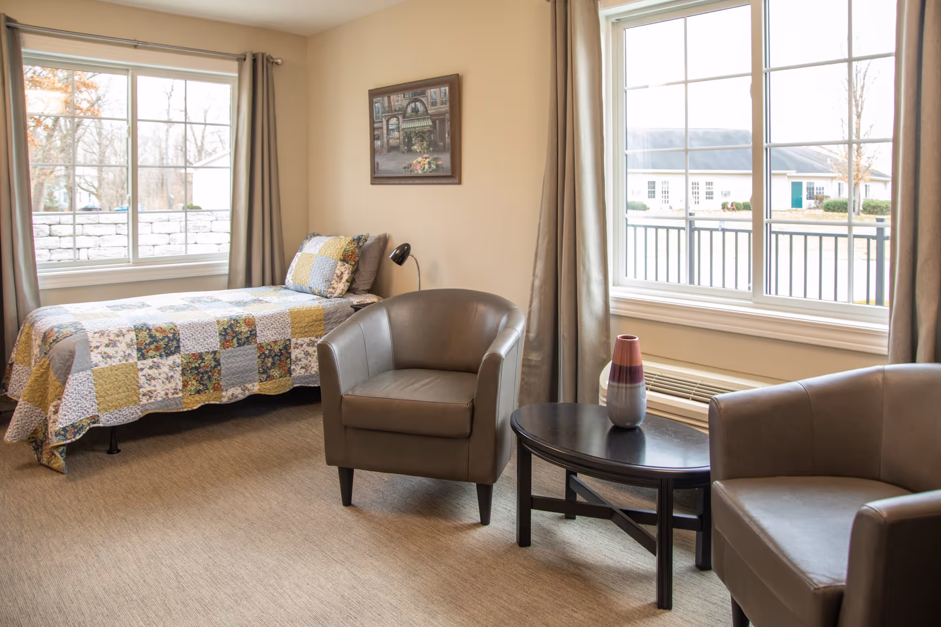 A cozy senior living bedroom with a single bed covered in a patchwork quilt, two gray armchairs, a black coffee table with a decorative vase, and large windows with beige curtains letting in natural light.