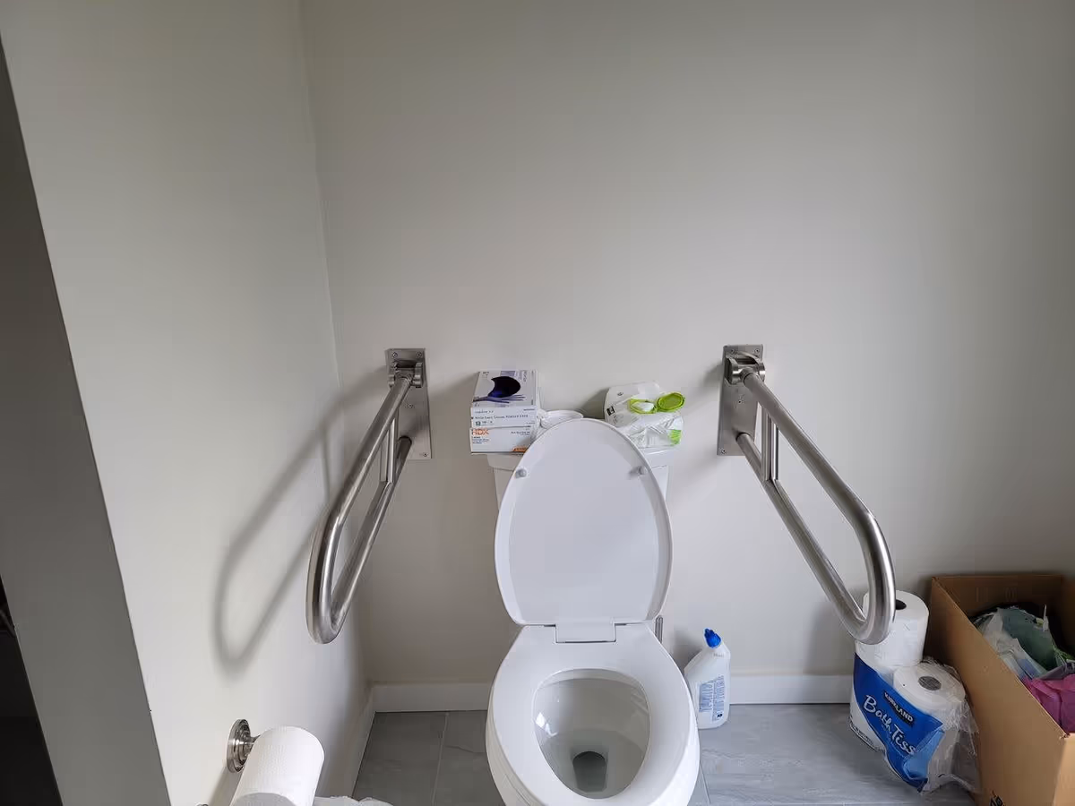 A bathroom with a white toilet that has the seat and lid raised. There are two stainless steel grab bars mounted on the walls on either side of the toilet. On top of the toilet tank, there are boxes of gloves and wipes. To the right of the toilet, there is a bottle of cleaning solution, a pack of toilet paper, and a cardboard box containing various items. A toilet paper roll is mounted on the left wall.