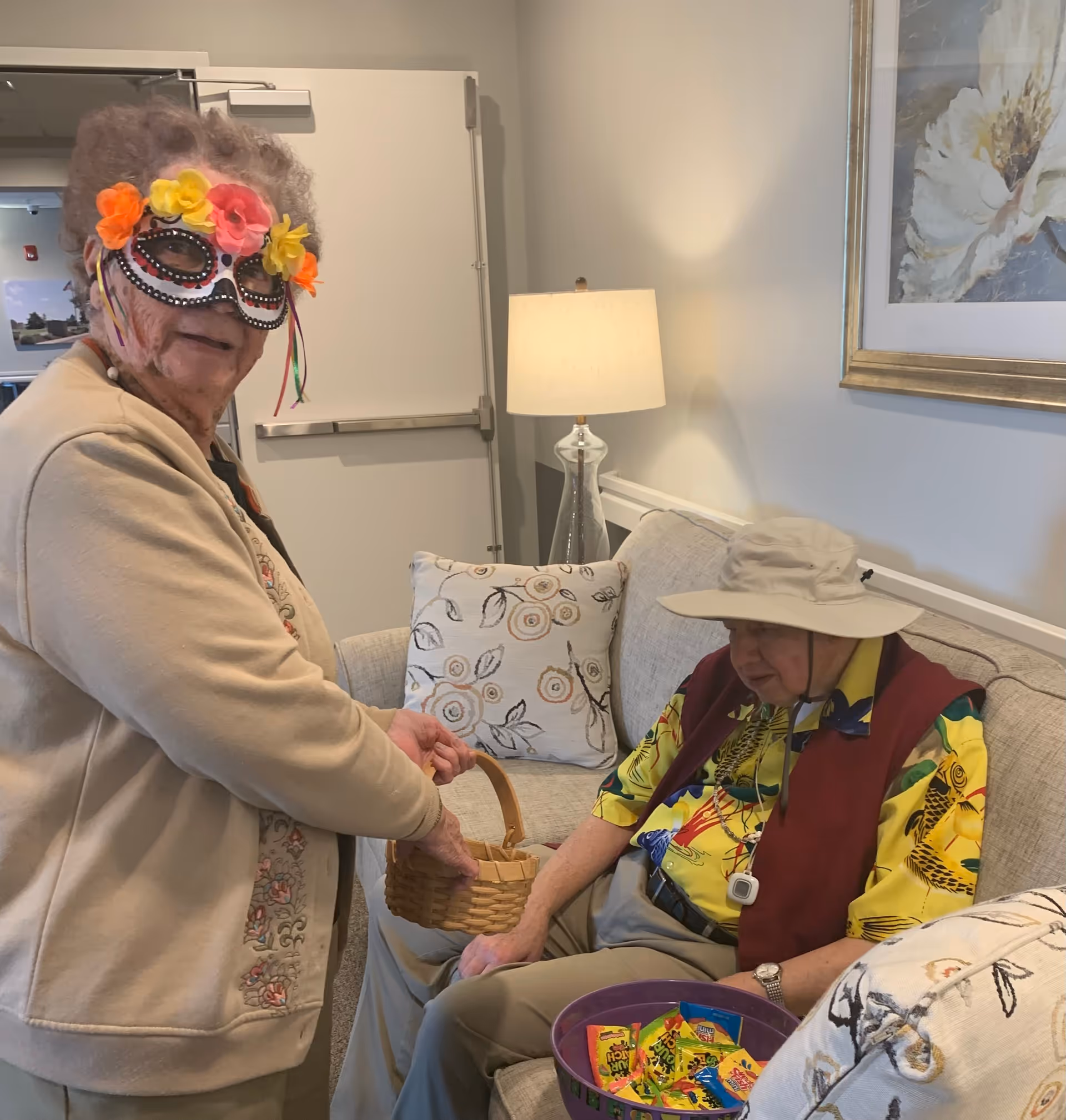 An elderly woman wearing a colorful floral mask and beige cardigan holds a small wicker basket while standing next to an elderly man seated on a couch. The man is wearing a wide-brimmed hat, a yellow patterned shirt, and a maroon vest, and holds a purple bowl filled with assorted candy. The setting is a cozy living room with a lamp, floral pillows, and a framed flower painting on the wall.