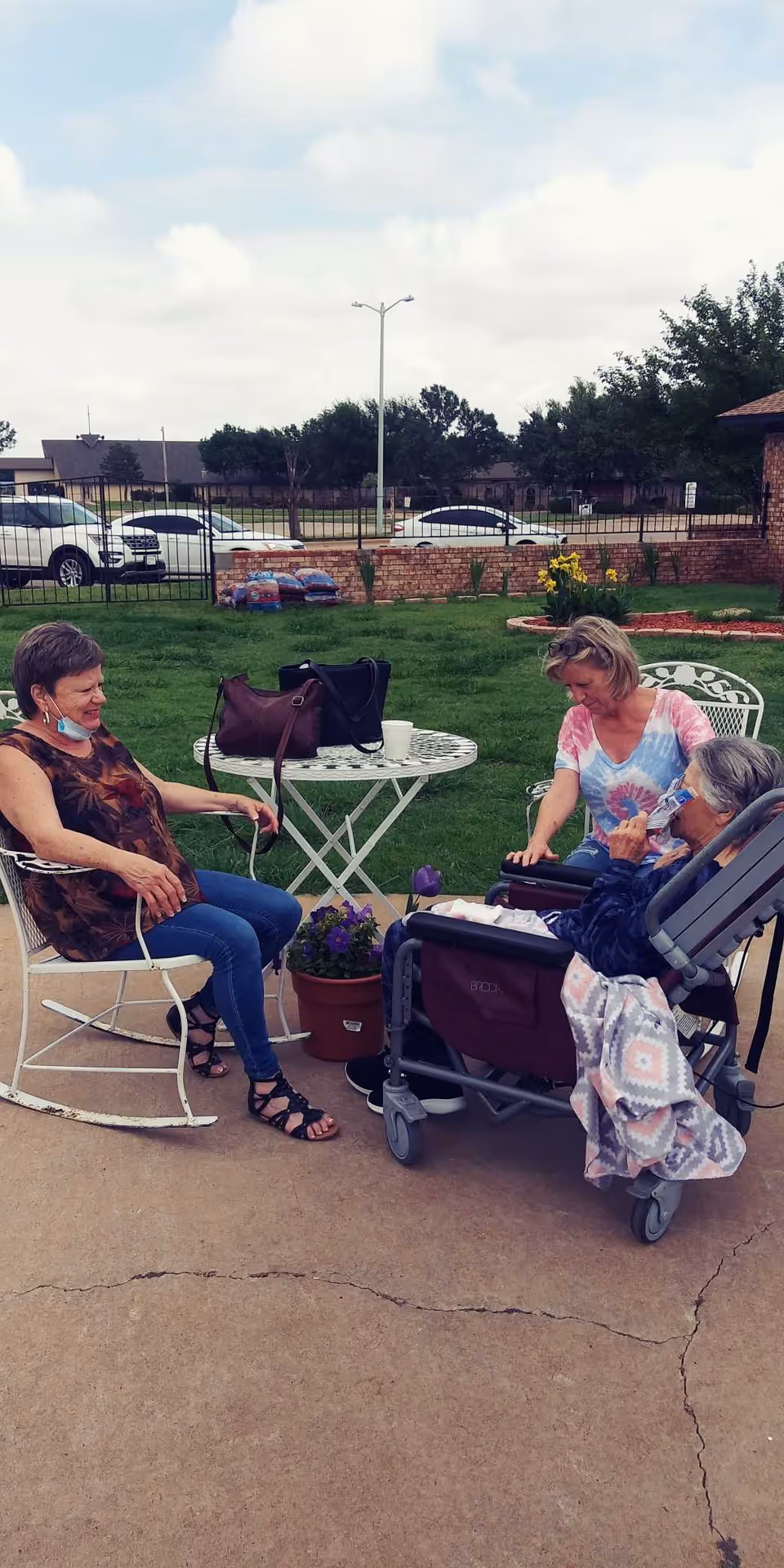 Three women sitting outside on a patio area with green grass and a brick wall in the background. One woman is seated in a wheelchair with a blanket, another woman is sitting on a white metal chair, and the third woman is leaning over the wheelchair. There is a white round metal table with a handbag and a cup on it, and a potted plant on the ground nearby. Cars and houses are visible in the background under a partly cloudy sky.