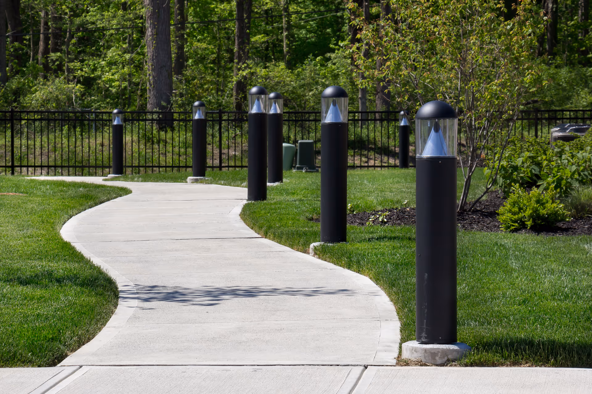 Curved concrete walkway lined with black bollard lights through a landscaped grassy area with trees in the background.