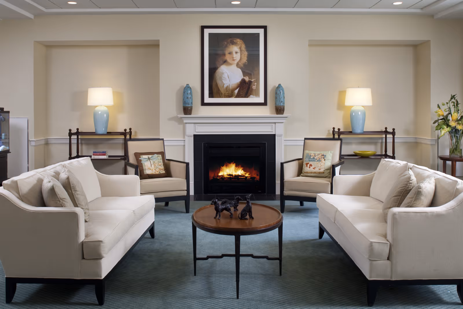A cozy living room with two beige sofas facing each other, two armchairs with decorative pillows, a round wooden coffee table with three small dog statues, a lit fireplace with a white mantel, and a framed portrait of a young girl above the fireplace. Two blue lamps on wooden side tables flank the fireplace, and a vase with yellow flowers is on a small table to the right.