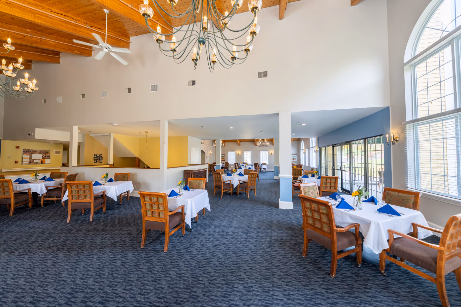 A spacious dining room with multiple tables covered in white tablecloths and set with blue napkins and small flower arrangements. The room features large windows allowing natural light, wooden ceiling beams, chandeliers, and carpeted flooring. Wooden chairs with cushioned seats surround the tables.