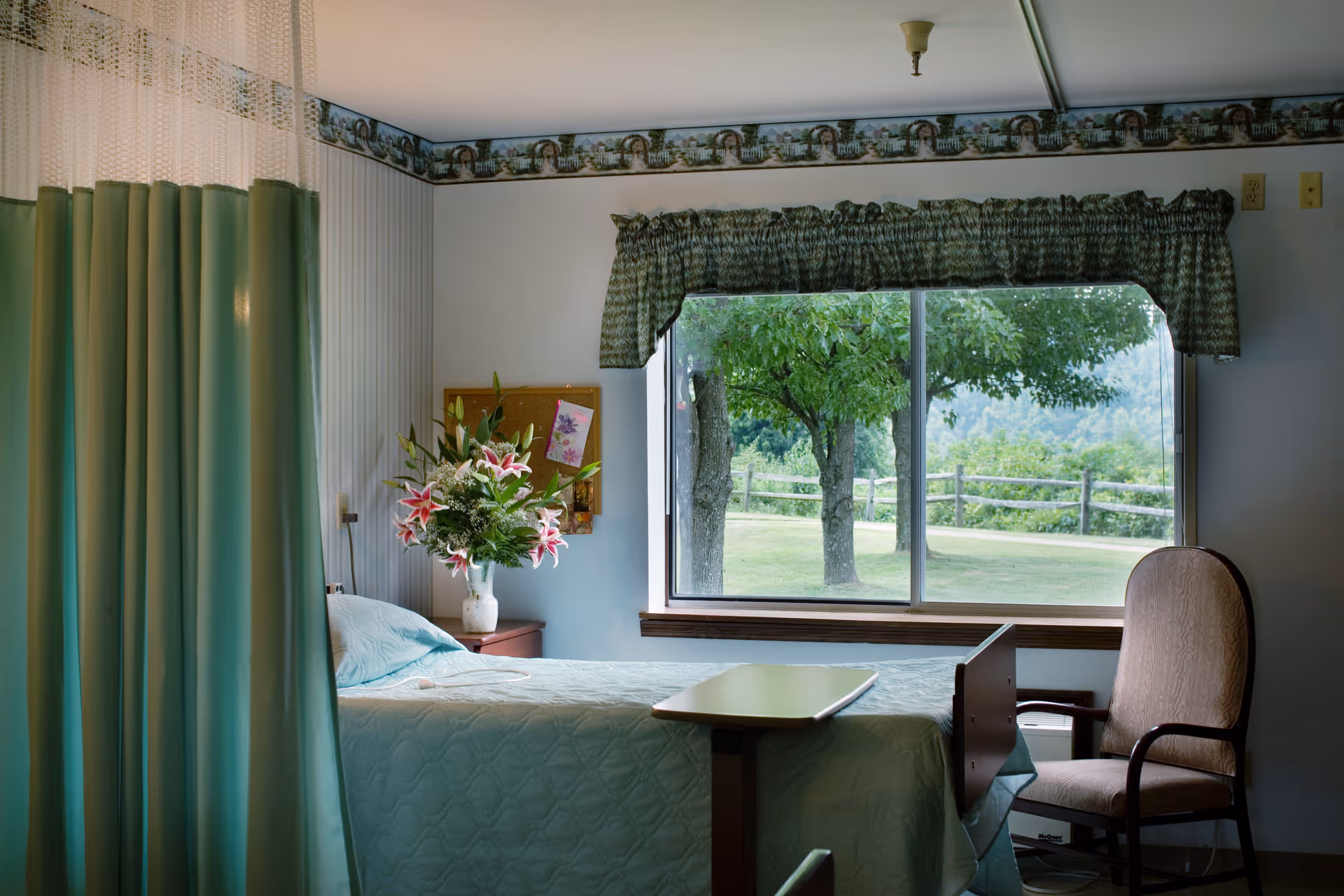 A sunlit senior bedroom with a bed, bedside table holding a vase of flowers, a chair and privacy curtain, and a large window looking out to trees and a fenced lawn.