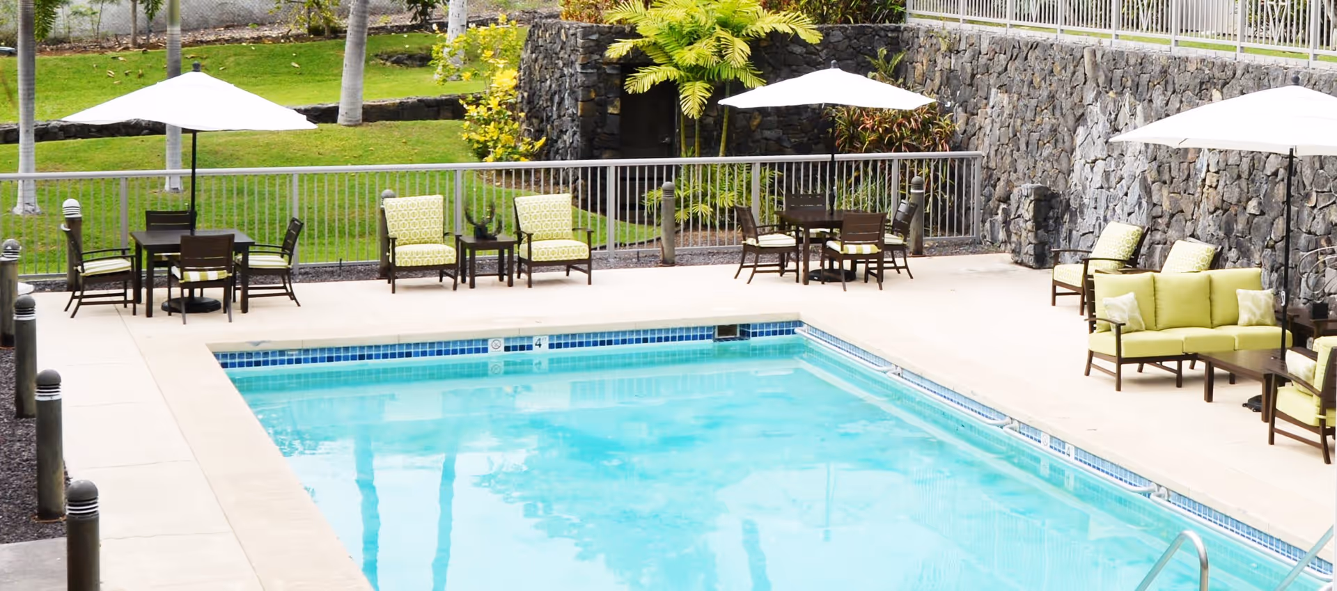 Outdoor swimming pool area with several seating arrangements including cushioned chairs, sofas, and tables with umbrellas. The pool is surrounded by a concrete deck and a stone wall with greenery and palm trees in the background.