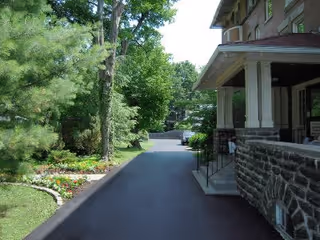 A paved driveway or pathway beside a building with stone and brick exterior. The path is lined with green trees and bushes on the left side, and there is a flower bed with colorful flowers near the edge of the path. The scene is outdoors with bright daylight.