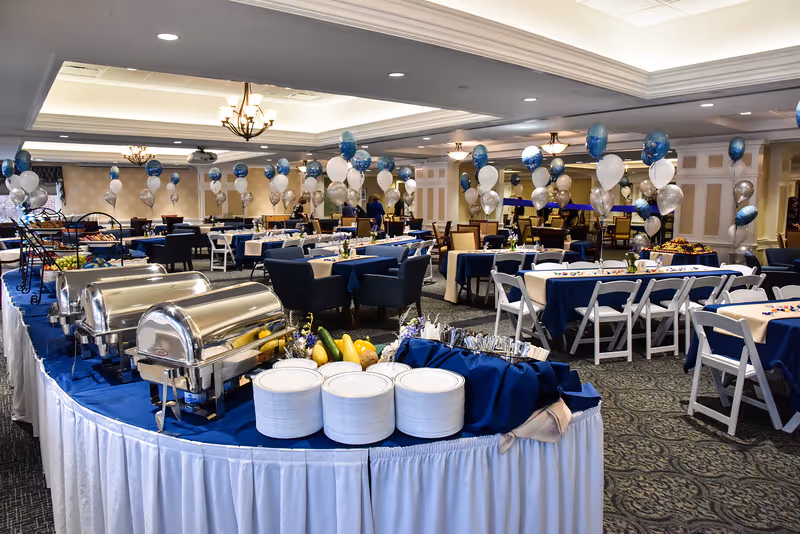 Large banquet dining room set up for an event with buffet chafing dishes, plates, tables draped in blue and white linens, and clusters of balloons.