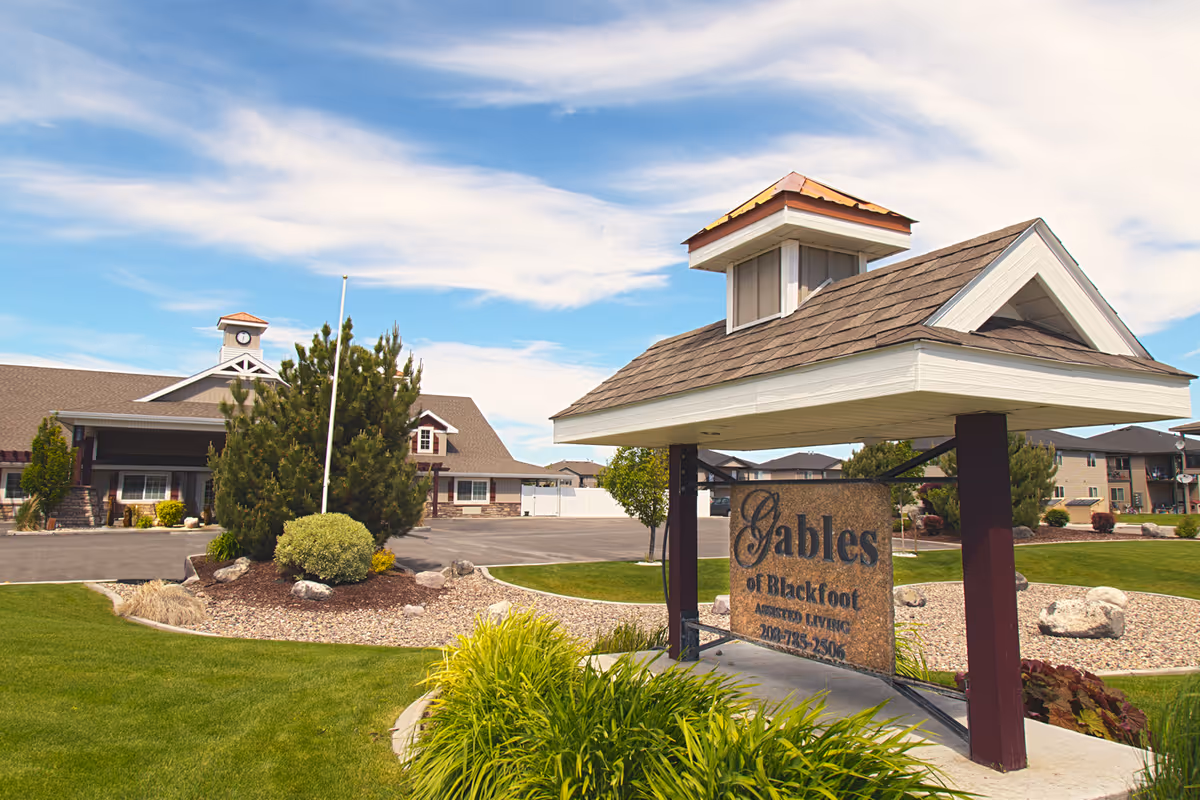 Exterior view of The Gables Assisted Living of Blackfoot facility with a landscaped lawn, a sign displaying the facility name, and a building with a clock tower under a partly cloudy sky.