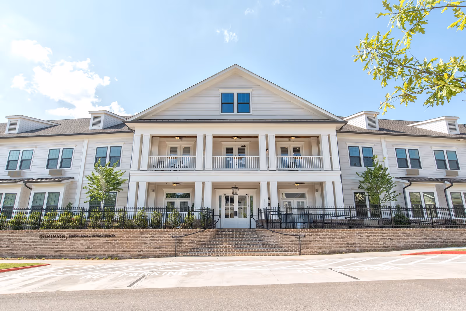 Front exterior of a two-story white senior living building with columns, covered balconies, stairs, and a brick retaining wall.