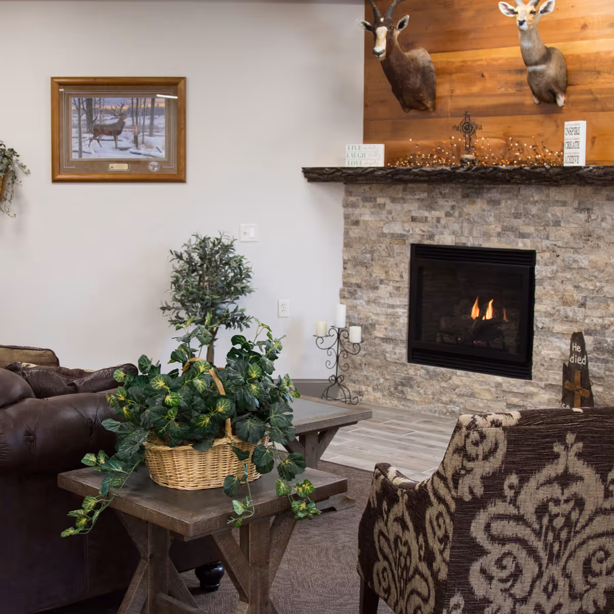 Cozy living room area with a stone fireplace featuring a lit fire. Above the fireplace are mounted deer heads and decorative items on the mantel. The room includes a patterned armchair, a brown leather sofa, a wooden side table with a basket of green leafy plants, and a framed picture of a deer on the wall.