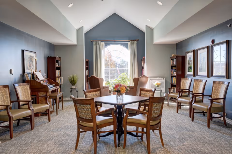 A bright and spacious common area with a high vaulted ceiling and a large arched window letting in natural light. The room features a square wooden table with four chairs in the center, two upholstered armchairs near the window, several wooden chairs with patterned cushions along the walls, bookshelves, framed artwork, and a piano on the left side. The walls are painted in a muted blue-gray color, and the floor is carpeted.