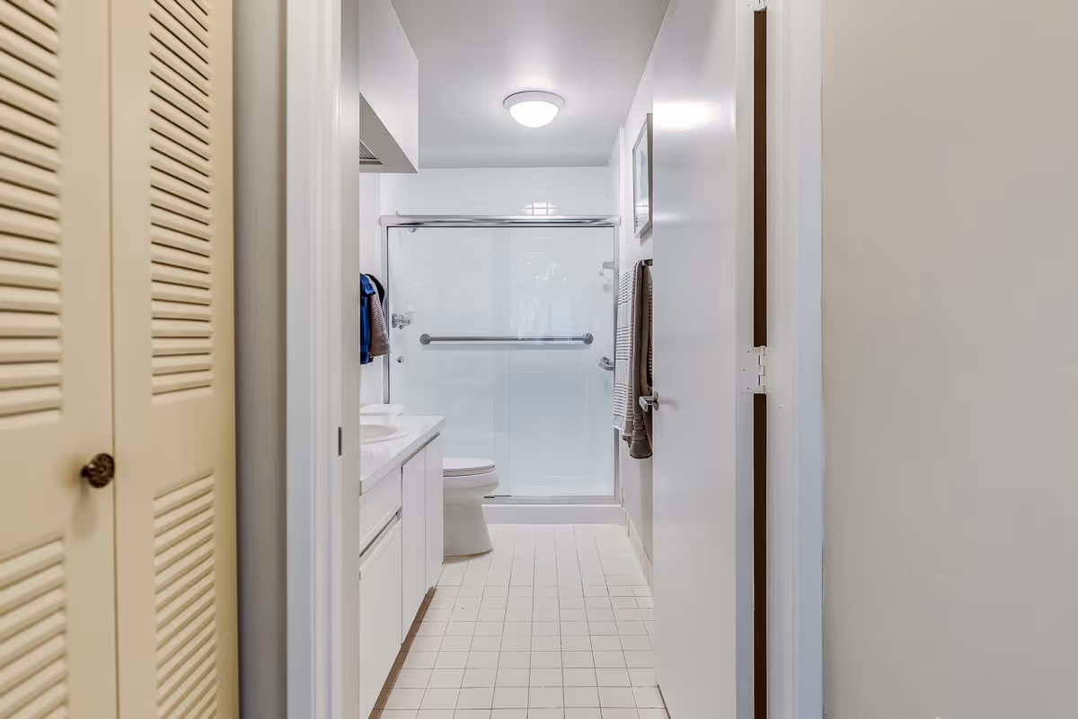 View down a short hallway into a clean white bathroom with a sink, toilet, and glass-enclosed shower.