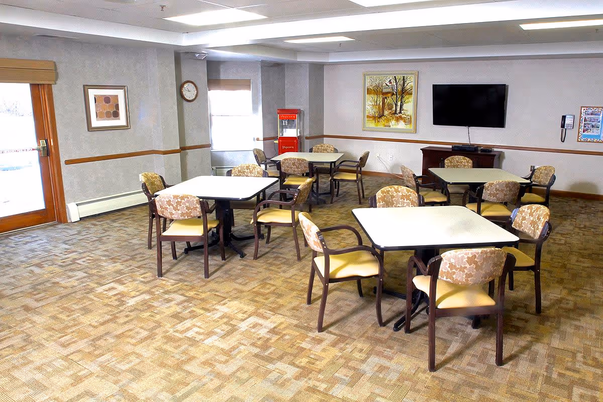 A senior living facility common dining area with several square tables and cushioned chairs arranged around them. The room has carpeted flooring, a wall-mounted TV, framed artwork, a clock, and a red popcorn machine near the windows. There is a door with a window on the left side letting in natural light.