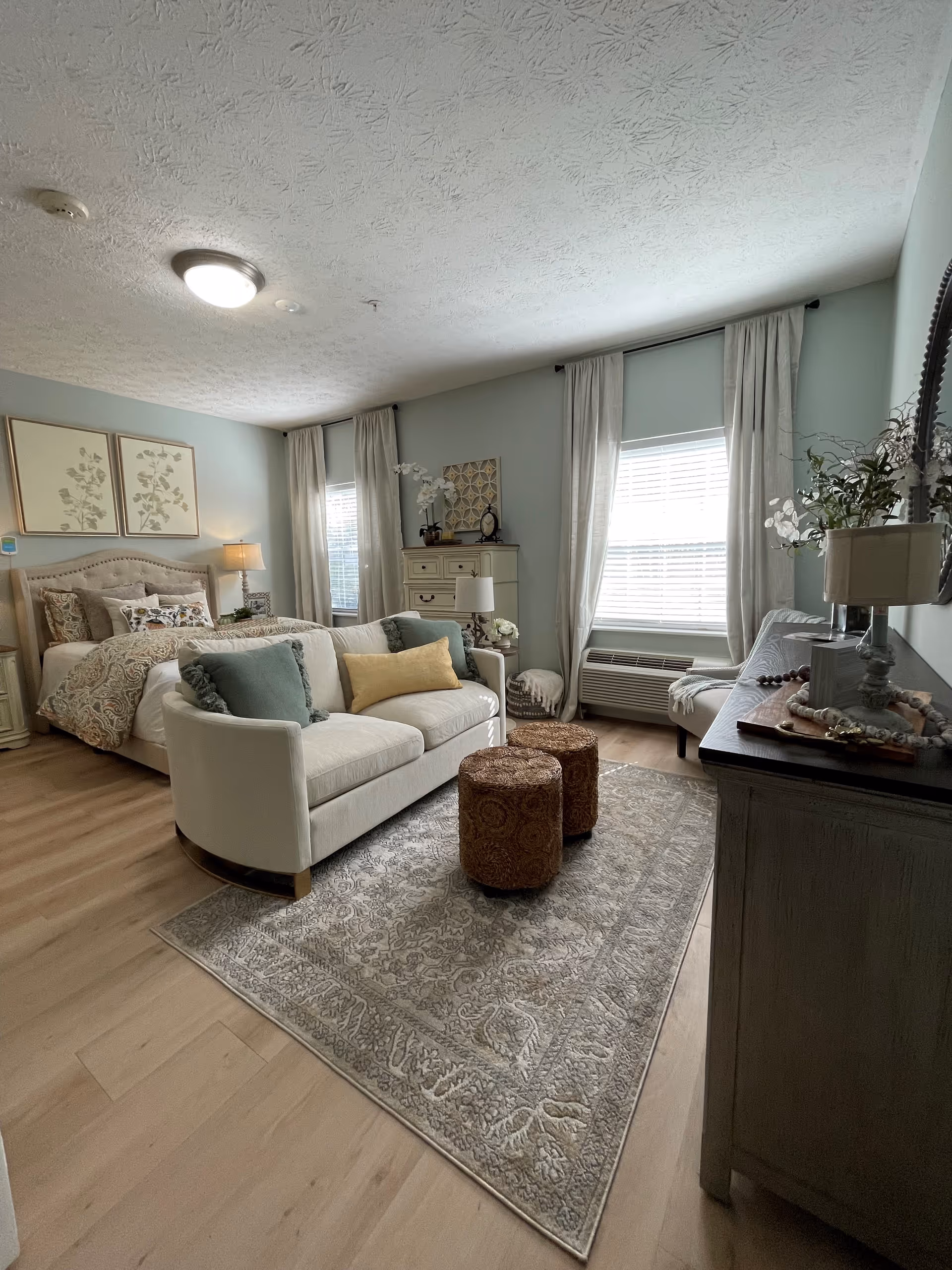 A cozy and well-decorated bedroom featuring a bed with patterned bedding and pillows, a small white sofa with cushions in front of the bed, two round ottomans on a patterned area rug, a dresser with decorative items, and two windows with white curtains allowing natural light to fill the room.