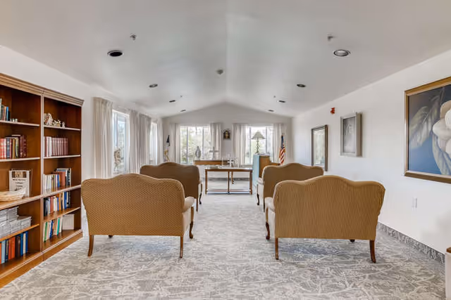 A bright and spacious common area in a senior living community featuring four upholstered armchairs arranged in two rows facing a wooden table near large windows with sheer curtains. The room has a patterned carpet, a wooden bookshelf filled with books and decorative items on the left, framed artwork on the walls, and an American flag near the window.
