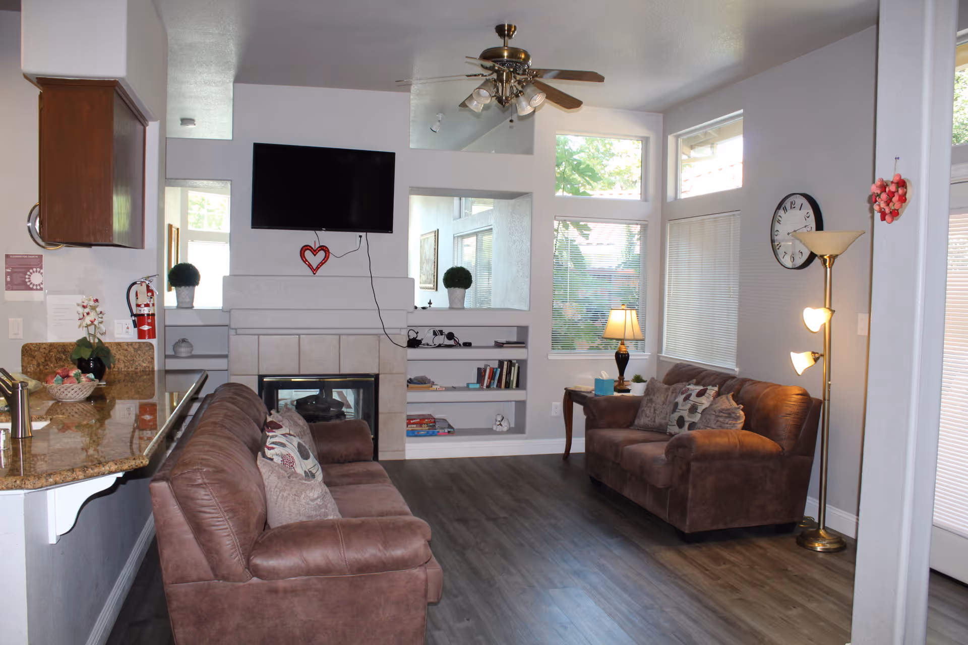 Cozy living room with two brown sofas facing a fireplace topped by a wall-mounted TV, built-in shelves, lamps, and a granite kitchen counter in the foreground.
