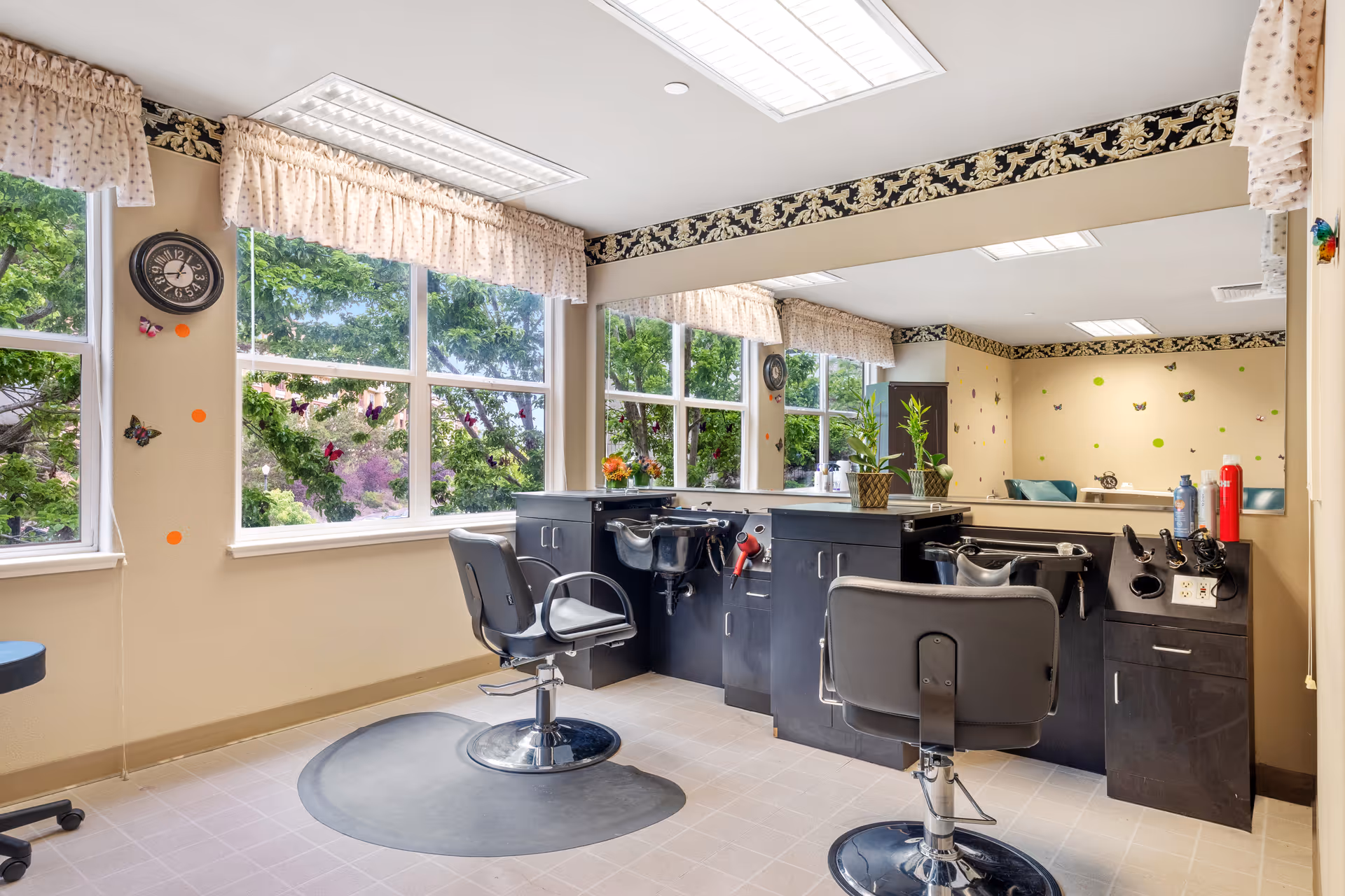 A bright and clean hair salon area inside a senior living facility with two black salon chairs in front of black counters equipped with sinks and hair styling tools. Large windows with floral valances let in natural light and show green trees outside. The walls are decorated with butterfly stickers and a clock is mounted on one wall.