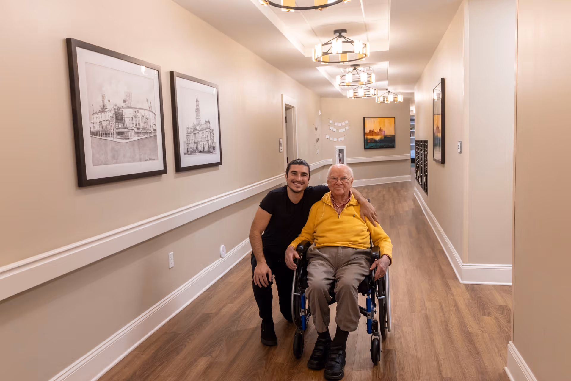 A young man kneeling and smiling next to an elderly man in a wheelchair in a well-lit hallway with wooden floors and framed artwork on the walls.