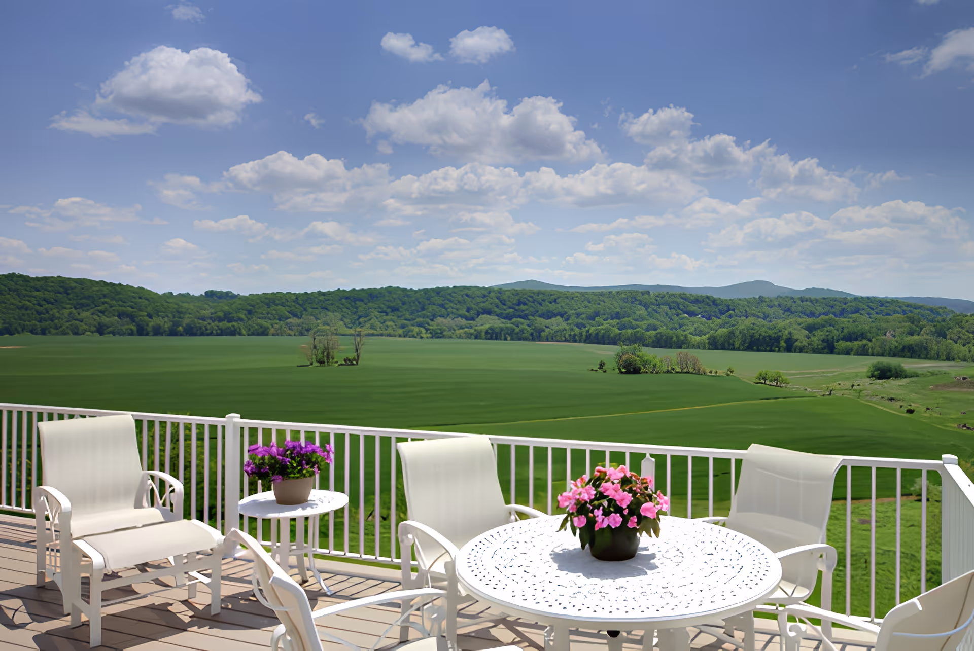 Outdoor patio area with white metal table and chairs, decorated with potted pink and purple flowers, overlooking a vast green field and forested hills under a partly cloudy blue sky.