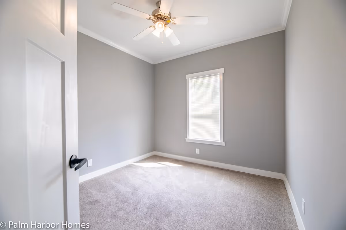 Empty room with gray walls, a window with blinds, beige carpet, a white ceiling fan with light, and a partially open white door with a black handle.