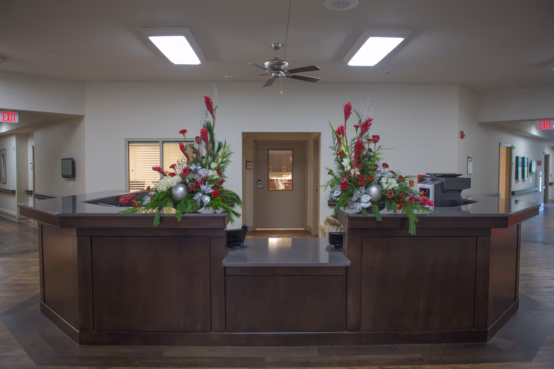 A dark wood reception desk decorated with two large floral arrangements in a bright hallway of a senior living facility.