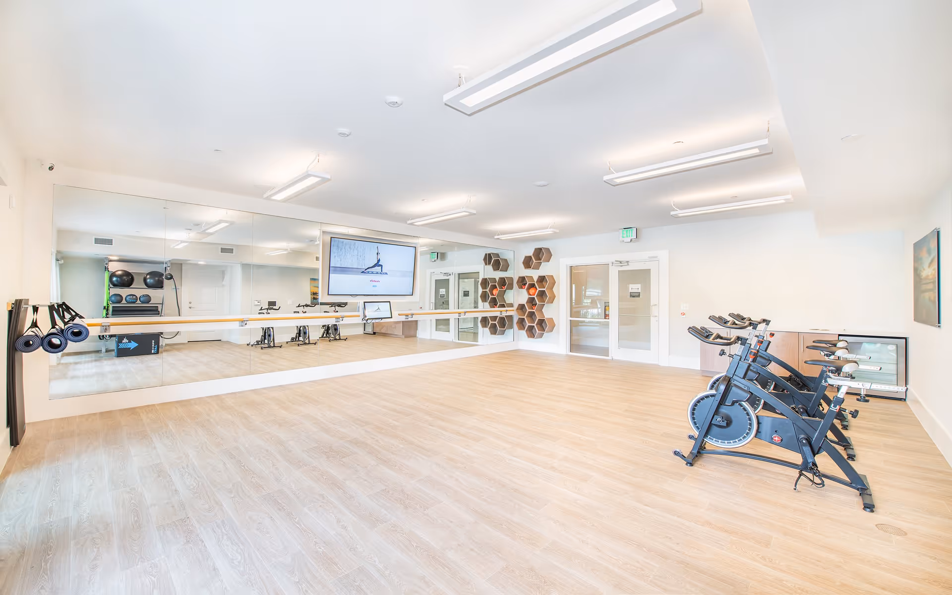 Bright and spacious exercise room with light wood flooring, a large wall mirror, mounted TV screen, exercise bikes lined up on the right, and shelves with exercise balls and equipment on the left.