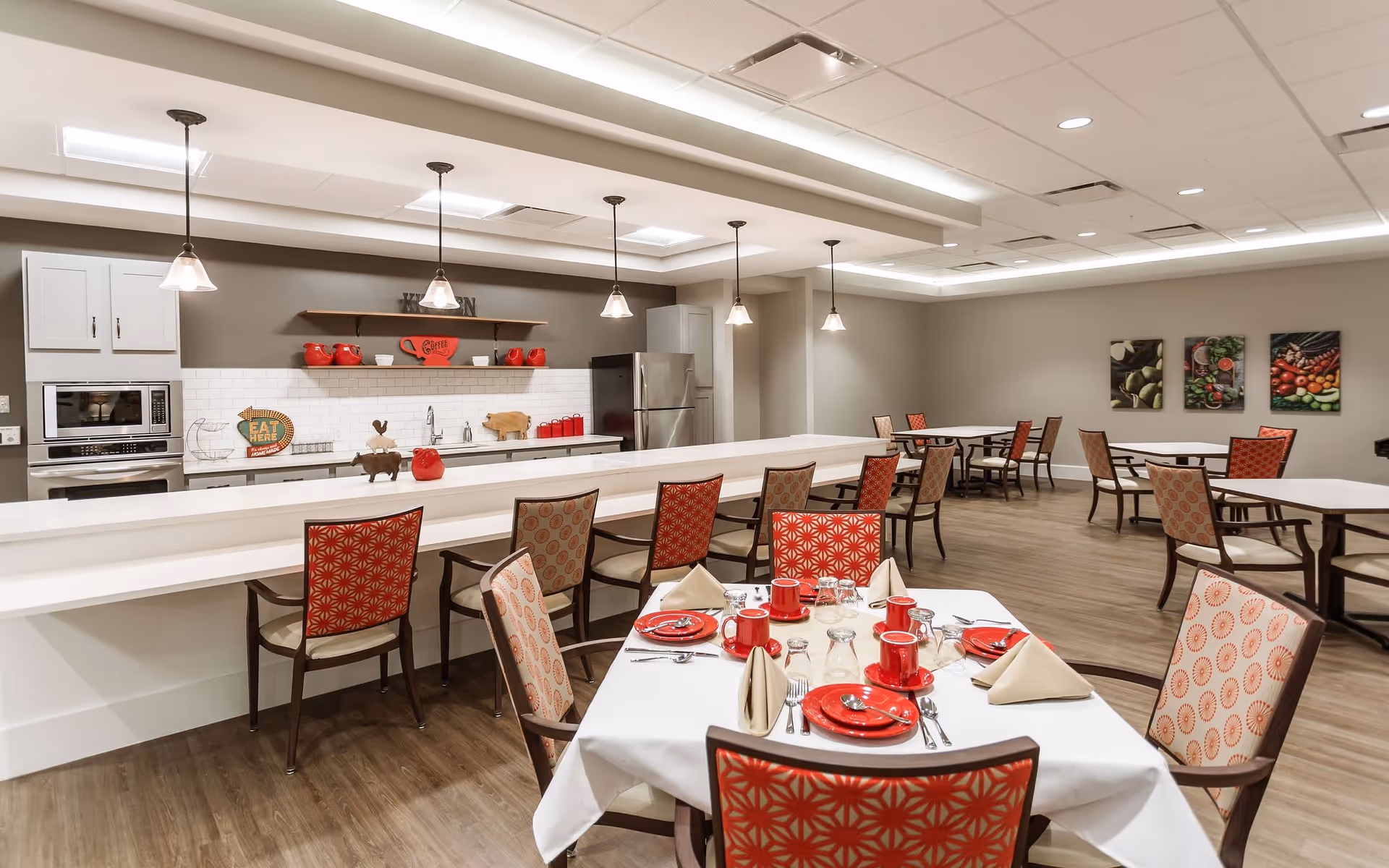 A spacious dining area with a modern kitchen in the background. The kitchen features white cabinets, a stainless steel refrigerator, microwave, and oven. There is a long counter with pendant lights hanging above and several chairs with red and beige patterned upholstery. In the foreground, a dining table is set with red plates, cups, silverware, and beige napkins. The room has wood flooring and neutral-colored walls with three framed pictures of vegetables on one wall.