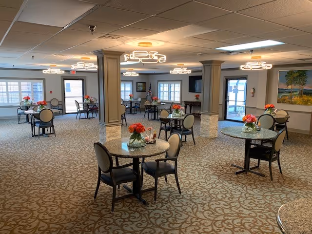 A spacious dining room with multiple round tables, each adorned with a vase of red and orange flowers. The room features beige patterned carpet, modern circular ceiling lights, and large windows with white shutters allowing natural light. There are several columns in the room, and paintings on the walls. Chairs with cushioned seats surround each table.