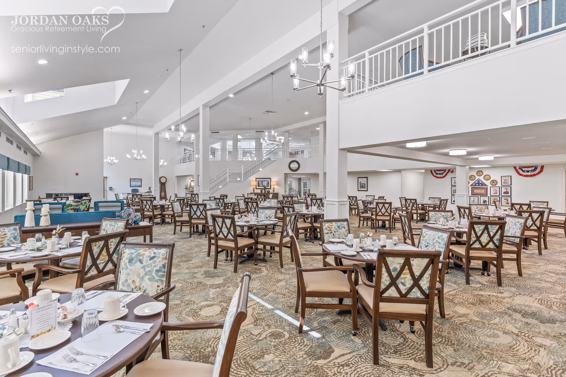 Large bright dining room with many set tables and chairs, chandeliers, and a mezzanine balcony.
