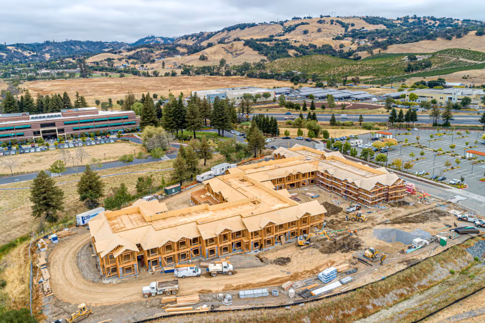 Aerial view of a large L-shaped wooden building under construction with construction vehicles, surrounding parking areas, roads, and hills in the background.