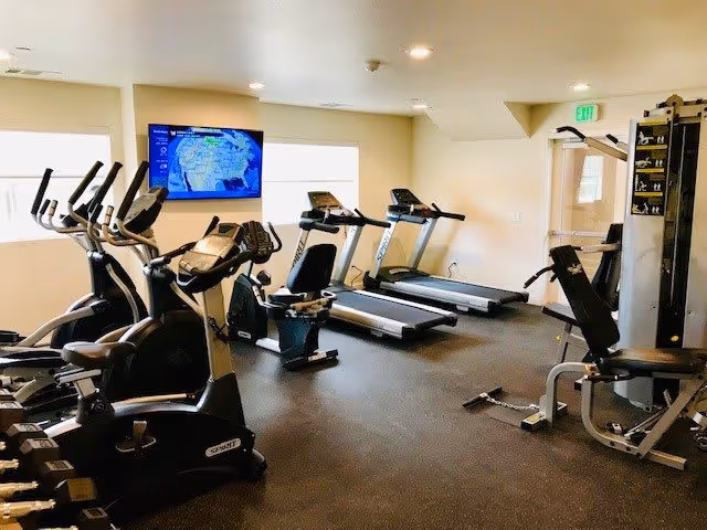 Interior view of a fitness room in a senior living community featuring exercise equipment including stationary bikes, treadmills, elliptical machines, and a multi-function weight machine. A wall-mounted TV screen displays a weather map. The room has beige walls, windows letting in natural light, and a rubberized floor suitable for workouts.