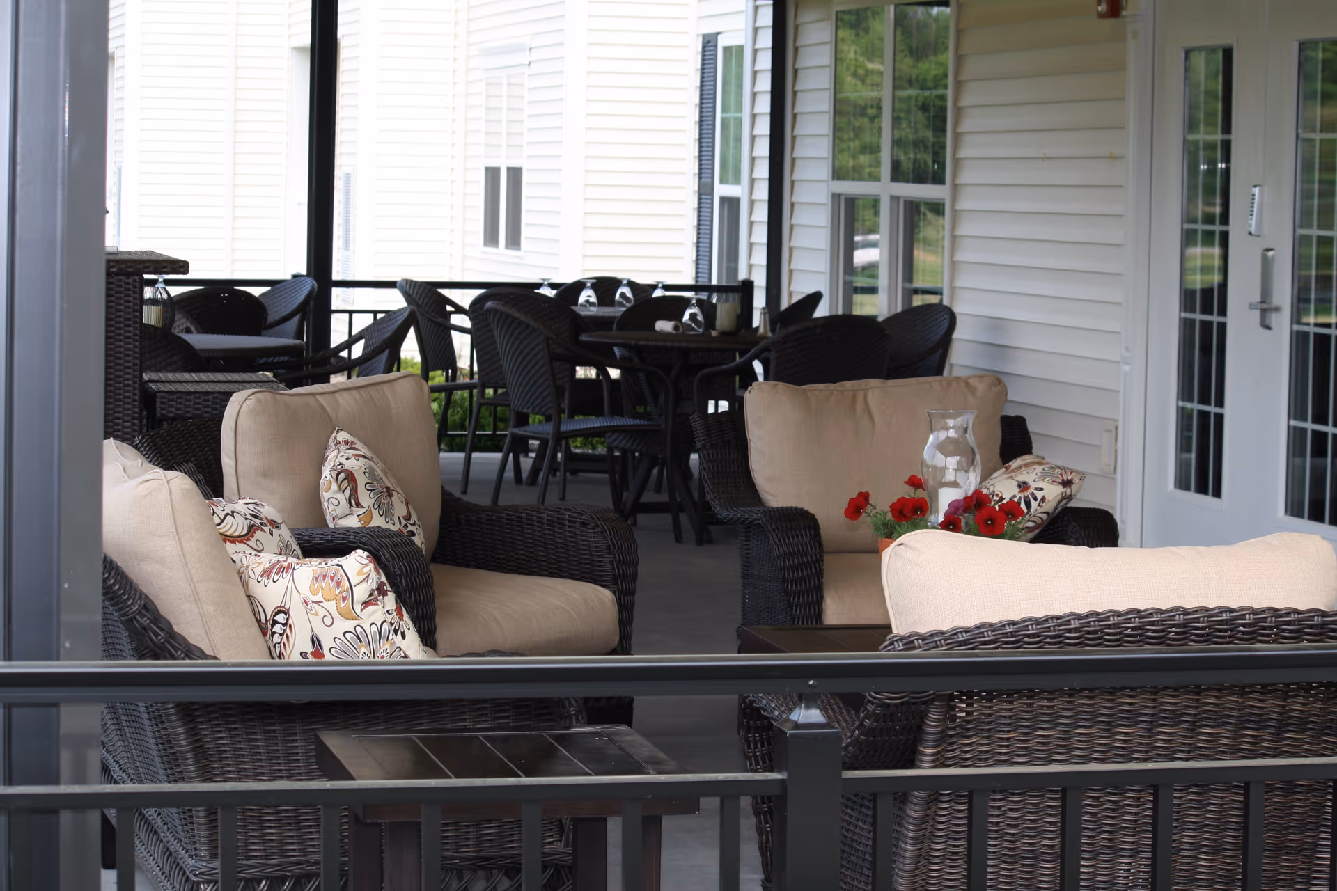 Covered outdoor patio area with wicker furniture including cushioned armchairs and a small table. There are decorative pillows on the chairs and a vase with red flowers on the table. In the background, there are additional tables and chairs near a white building with windows and a door.
