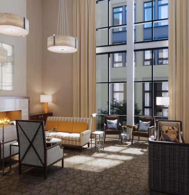 A bright and spacious living room area with large floor-to-ceiling windows letting in natural light. The room features a fireplace, a striped sofa with a yellow cushion, several armchairs with cushions, two modern pendant lights hanging from the ceiling, and beige curtains framing the windows.