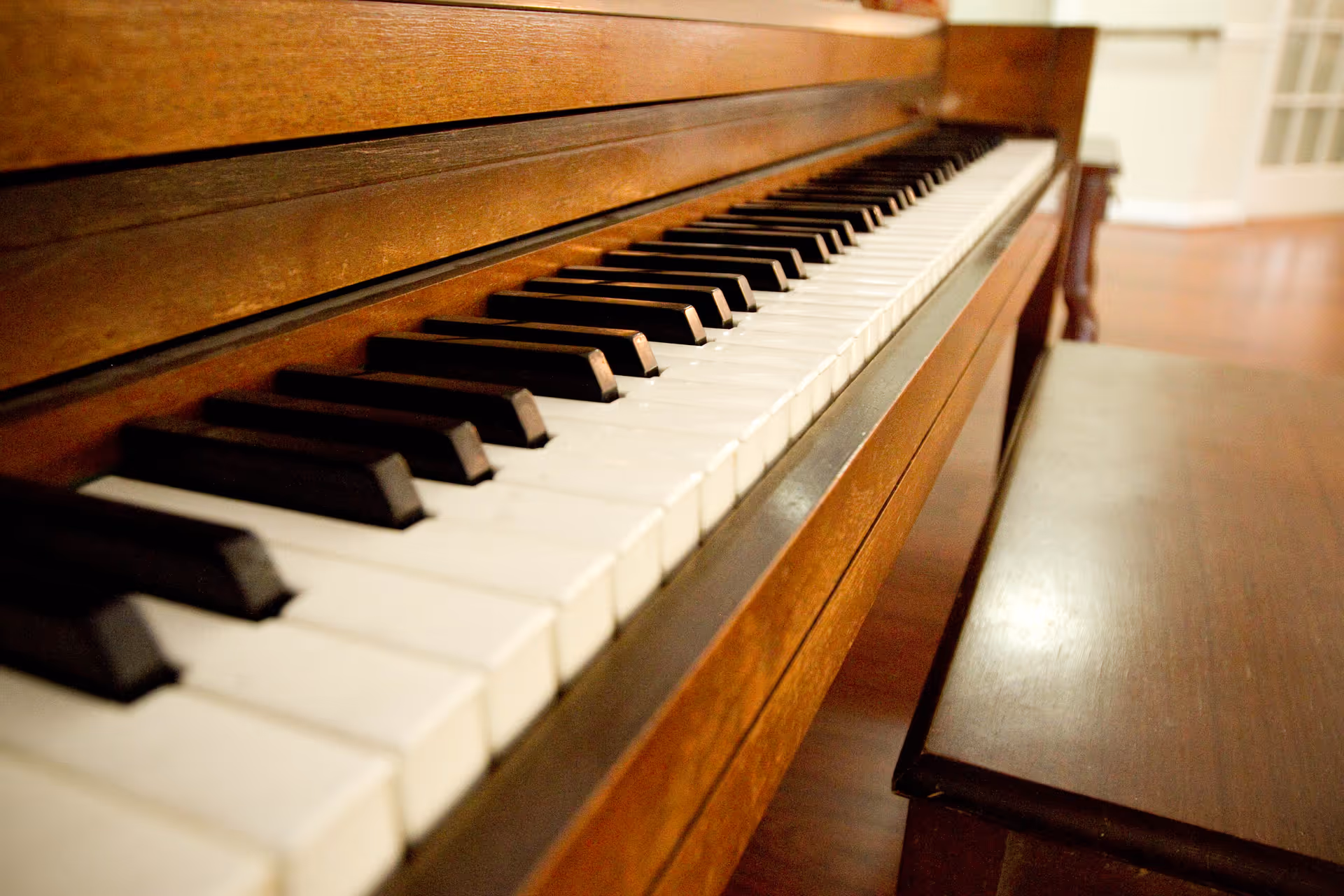 Close-up view of the keyboard of a wooden piano with a matching wooden bench beside it, set in an indoor space with wooden flooring.