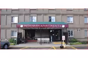 Exterior view of the entrance to Faith Haven Apartments, showing a covered entryway with a maroon awning displaying the facility name. The building is a multi-story structure with several windows, and there is a bench and some potted plants near the entrance.