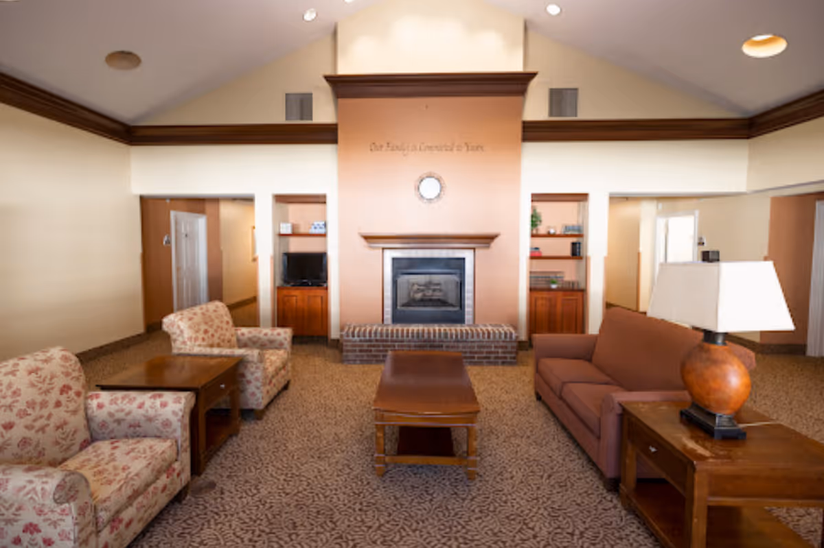 A cozy senior living facility common area with a brick fireplace centered on a peach-colored wall. Above the fireplace is a round clock and the phrase 'Our Family at [illegible] Years'. The room features two floral-patterned armchairs on the left, a brown sofa on the right, wooden coffee and side tables, and a lamp on the right side table. Built-in shelves and cabinets flank the fireplace, with a small TV on the left side. The ceiling is vaulted with recessed lighting.