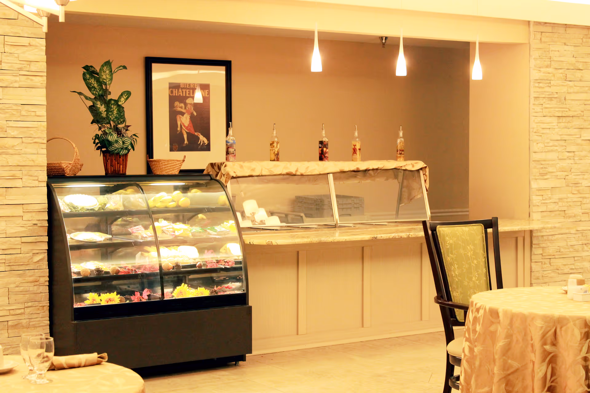 Interior view of a dining area with a glass display case filled with food items, a serving counter with bottles on top, a framed picture on the wall, and a table with a beige tablecloth and a green cushioned chair.