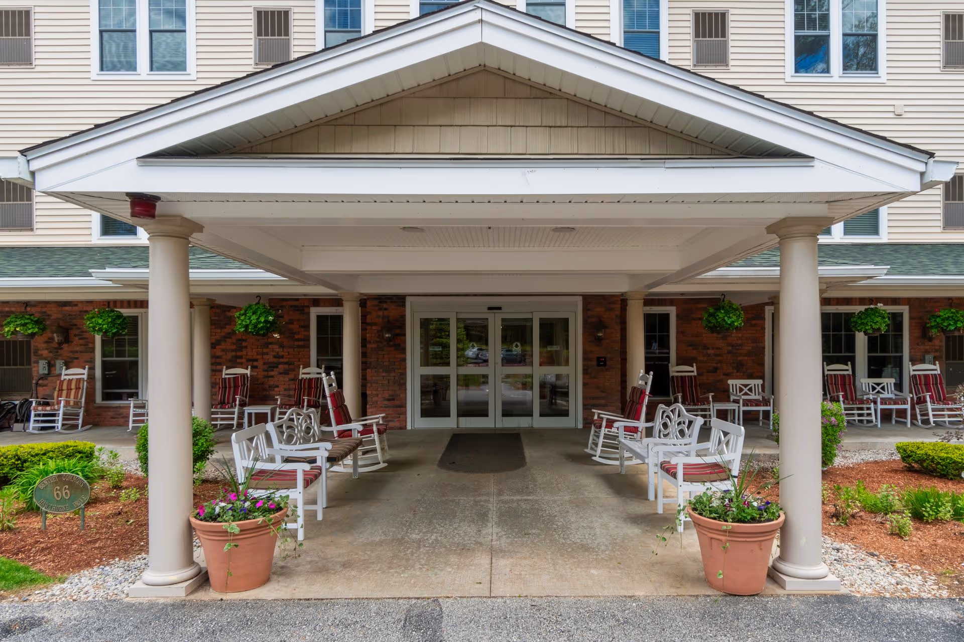 Entrance of Bentley Commons at Bedford featuring a covered porch with white columns, multiple white rocking chairs with red cushions, hanging green plants, potted flowers, and a brick wall background with windows and air conditioning units.