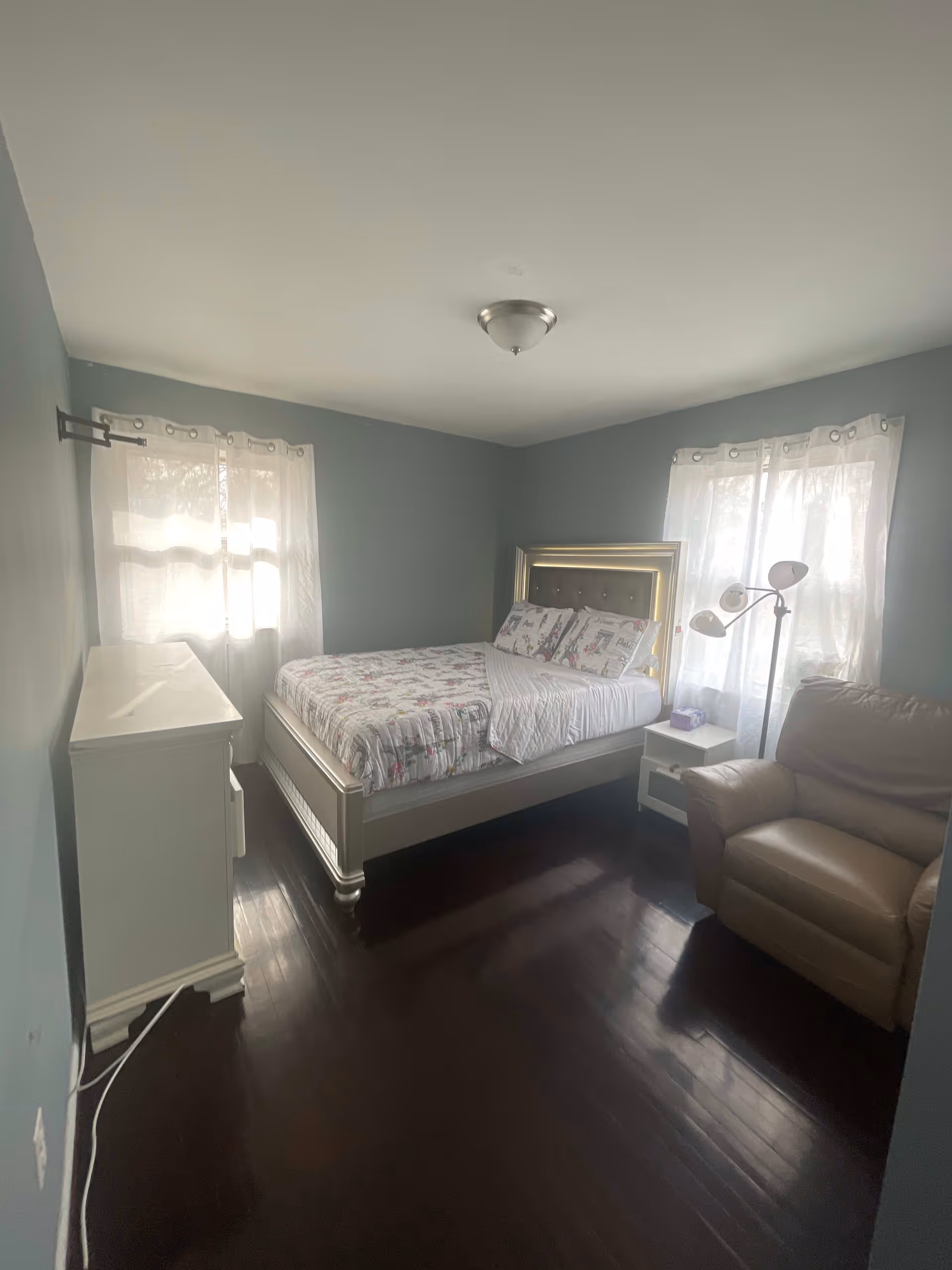 A bedroom with light blue walls and dark wooden floor. The room contains a bed with a decorative headboard and floral bedding, a white dresser, a white nightstand with a box of tissues, a beige armchair, and a floor lamp with three adjustable lights. Two windows with sheer white curtains allow natural light into the room.