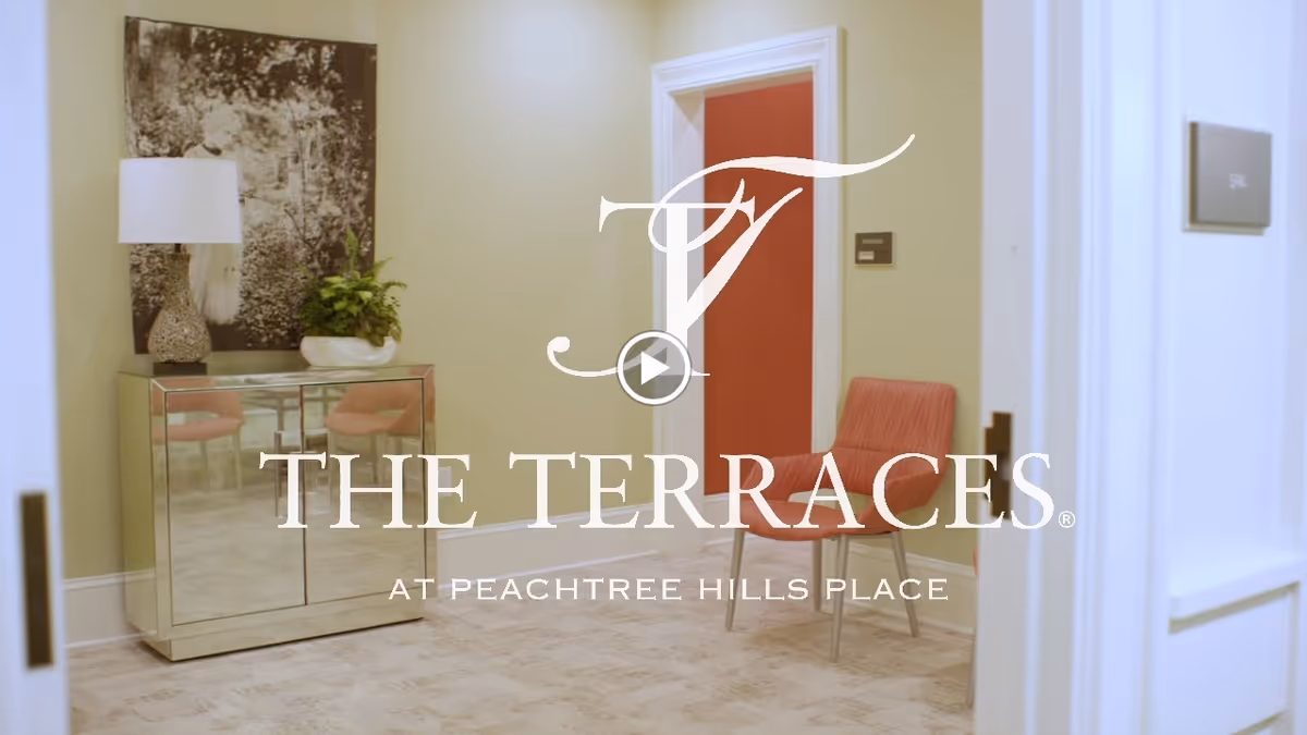 A decorated interior hallway with a mirrored cabinet, table lamp and plant, a red chair by a doorway, and a large overlaid facility logo.