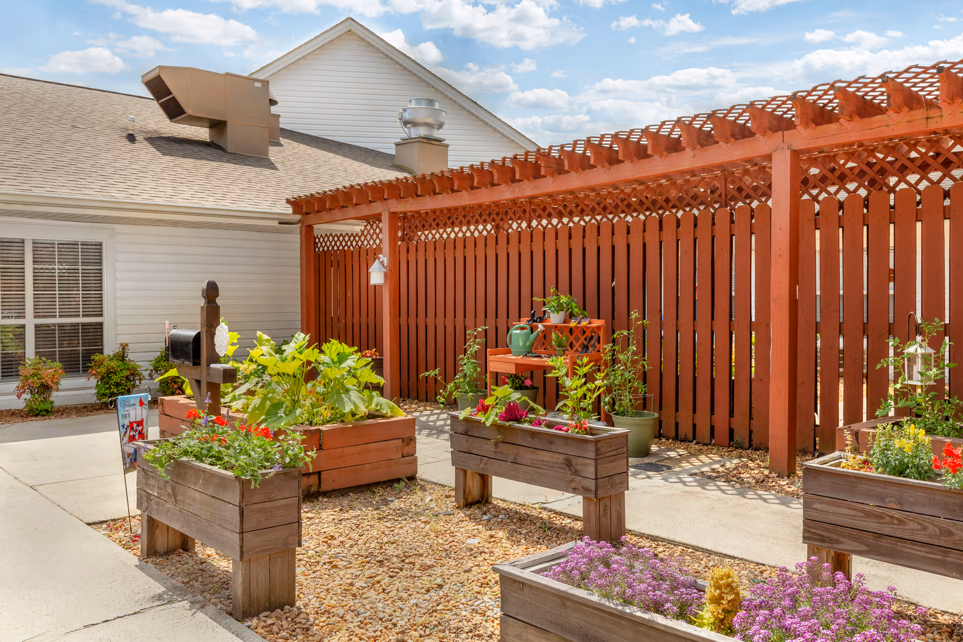 Sunlit outdoor courtyard with raised wooden planter boxes filled with flowers and a red wooden pergola beside the building.