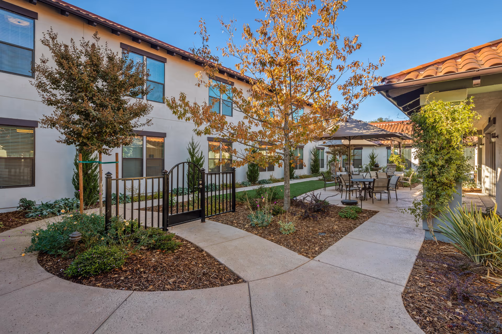 Outdoor courtyard area at Oakmont of East Sacramento featuring a paved walkway, small trees with autumn leaves, landscaped garden beds, and a seating area with tables, chairs, and umbrellas. The courtyard is surrounded by a two-story building with windows and a tiled roof.