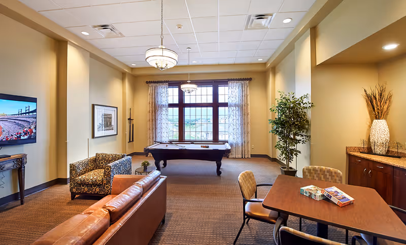 A senior living facility common area with a pool table near large windows with sheer curtains. The room has a brown leather sofa, a patterned armchair, a wooden table with four chairs, and a flat-screen TV mounted on the wall. There is also a cabinet with a decorative vase and plant in the corner.