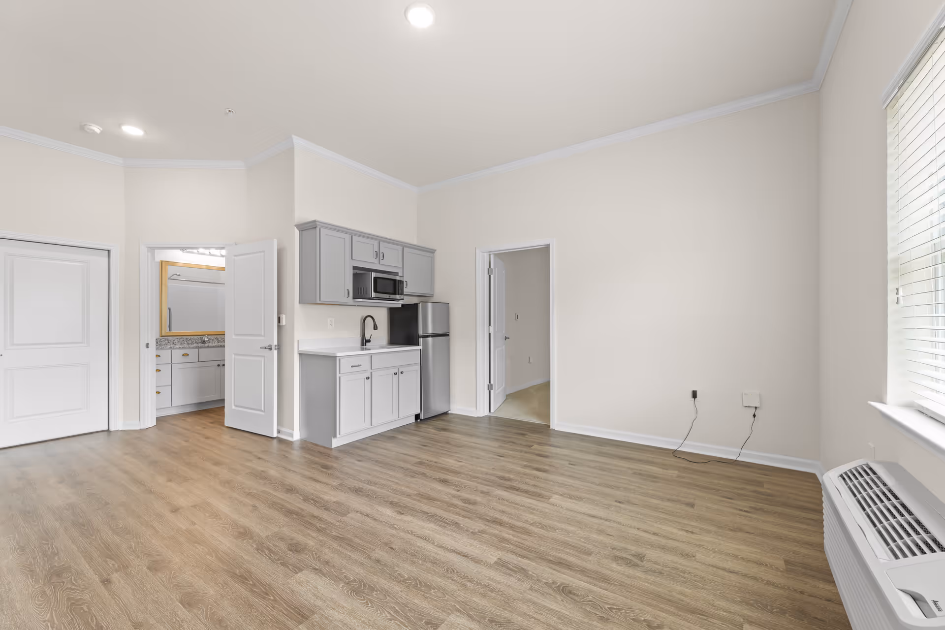 Empty studio-style living area with a small kitchenette, wood-look flooring, and an open doorway to a bathroom.