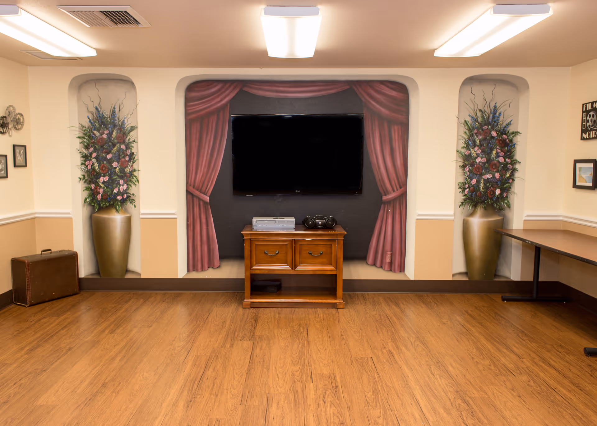 A communal room with a wall-mounted TV centered between painted curtain murals, a wooden console beneath it, floral mural niches on either side, and a hardwood floor.