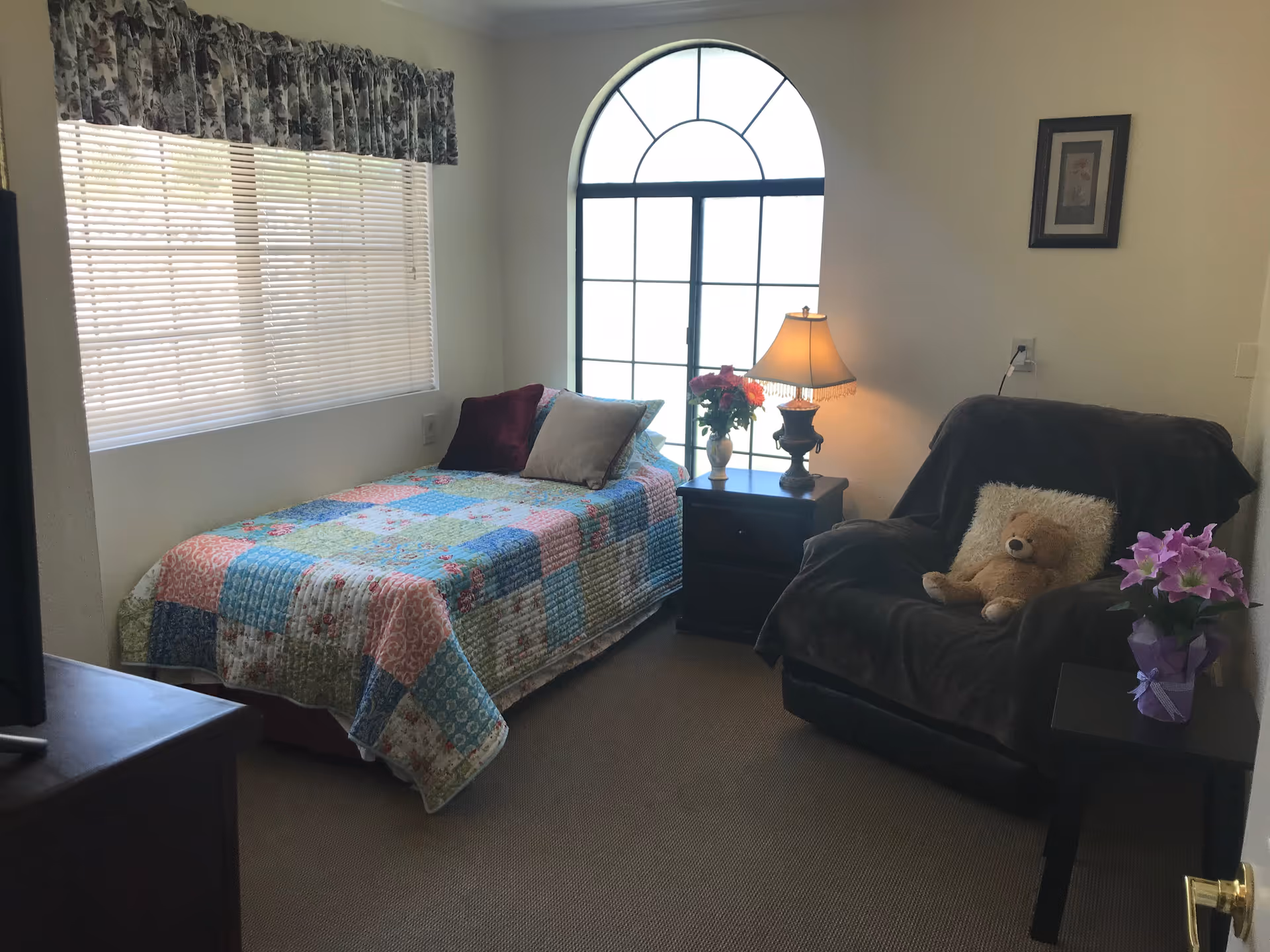 A cozy bedroom with a single bed covered in a colorful patchwork quilt, two pillows, a nightstand with a lamp and a vase of flowers, a large arched window, a comfortable armchair with a teddy bear and a fluffy pillow, and a small table with a potted plant.