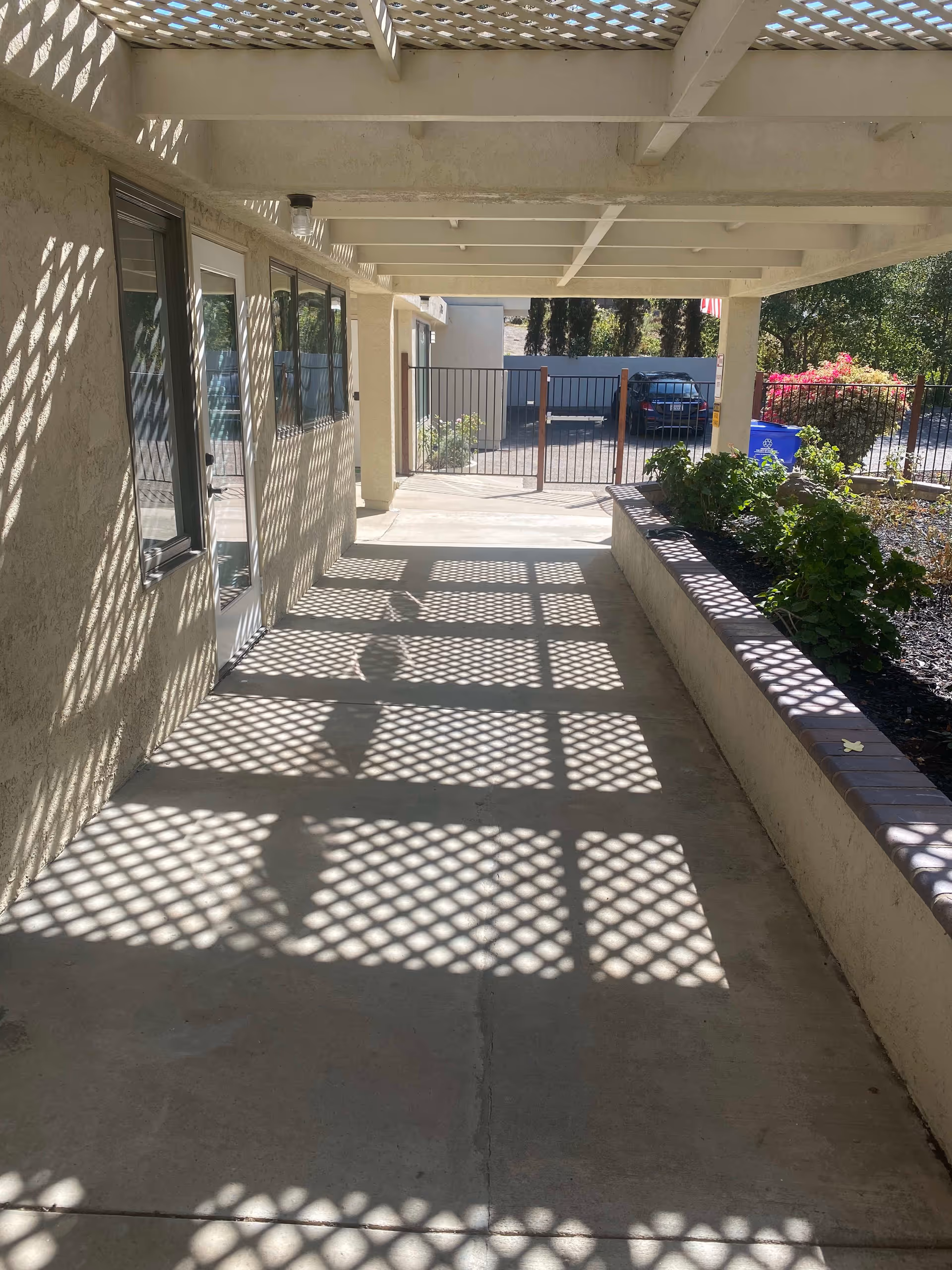 Covered outdoor walkway with lattice roof casting patterned shadows on the concrete floor, adjacent to a building wall with windows and a door, and a raised garden bed with plants on the right side. A metal gate and parked car are visible in the background.