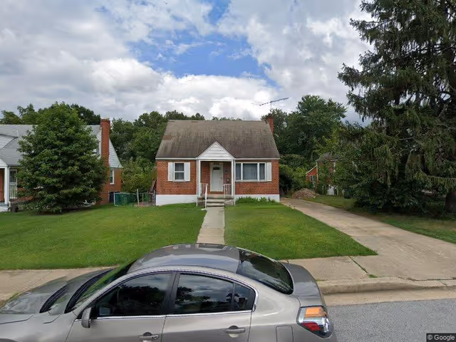 Front view of a small brick single-family house with a lawn, driveway, and a car parked in the street.