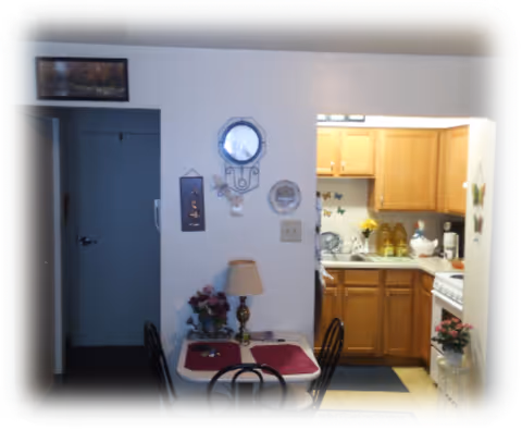 Small dining area with a table covered by two red placemats and surrounded by black metal chairs. A lamp and a flower arrangement are on the table. The dining area is adjacent to a compact kitchen with wooden cabinets, a white stove, and a sink. The wall above the table features a clock, a decorative plate, and some wall hangings. A doorway leads to another room or hallway.
